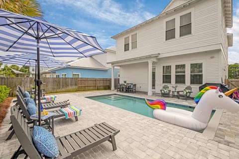 Spacious backyard oasis features a sparkling pool, loungers under striped umbrellas, and a covered patio—ideal for sunny afternoons and poolside relaxation.
