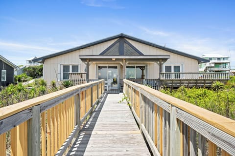 Charming beachfront rental with elevated boardwalk entrance, surrounded by coastal dunes and native vegetation under clear coastal skies.