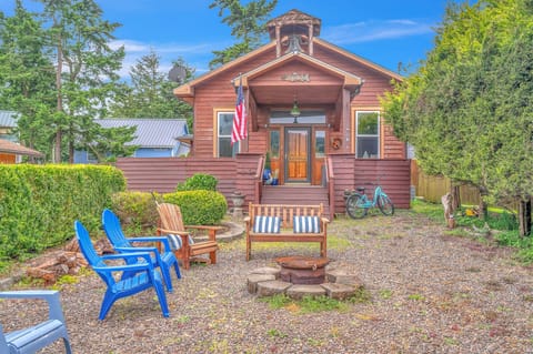 Charming red cottage with a welcoming front porch and fire pit seating area, surrounded by mature trees in a peaceful neighborhood setting.
