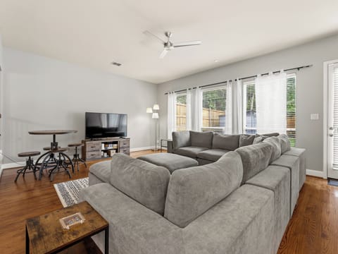 Sink into the plush sectional and relax in your bright, airy living space—complete with a flat-screen TV, warm hardwood floors, and peaceful garden views through sheer curtains.