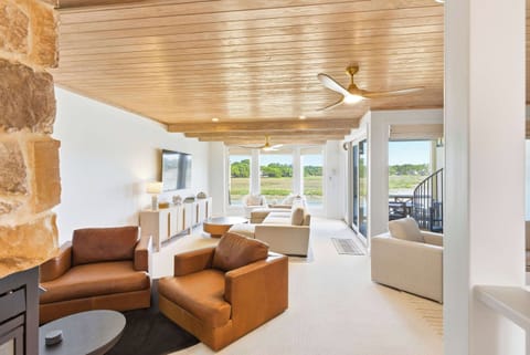Grand living room with soaring wood-plank ceiling, dramatic stone fireplace, rich leather armchairs, and sweeping waterfront views through the full-wall sliders.