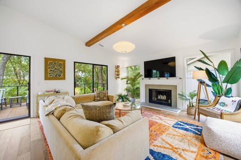 Velvet cushions and a vibrant patterned rug anchor this airy living room. A honey-toned wooden beam spans the ceiling, while glass doors draw the surrounding leafy forest right into your morning coffee spot.