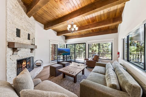 Living room with vaulted wood ceilings, stone fireplace, gray sofa, and large windows overlooking trees and deck.
