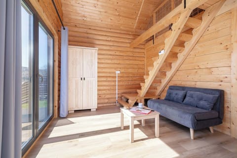 A bright living room with large windows, featuring a gray sofa and a wooden staircase leading to the upper floor. The room is bathed in sunlight thanks to the wide patio doors.