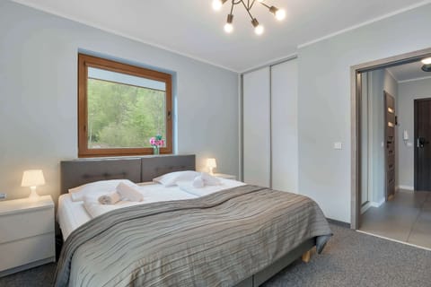 A cozy bedroom features a large double bed with crisp white linens and a plush gray headboard. Natural light streams in through a window, illuminating the neutral-toned walls and a nearby doorway.