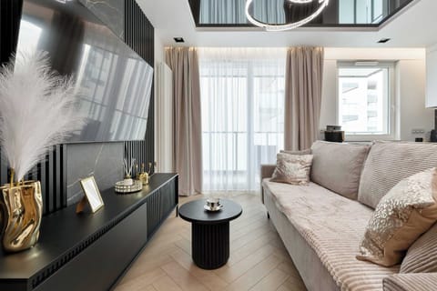 A modern living room features a dark media console and a beige textured sofa. Large windows draped with light-colored curtains brighten the space, which is accented by a small round coffee table.