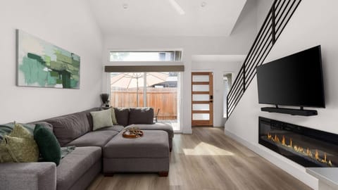 Light-filled living room with a plush sectional, a large sliding glass door opening directly to the outdoor patio, and a modern open staircase — a bright, airy layout that flows effortlessly between indoors and out.