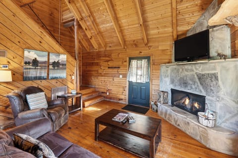 Warm cabin living room with a glowing stone fireplace, a queen sleeper sofa, and sunlight pouring through big mountain‑view windows. (Maggie's Cabin)