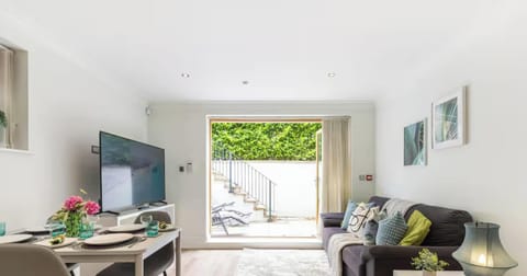 Relax in this bright, open-plan living and dining area, featuring a dark sofa accented with colorful pillows, a set dining table, a large flat-screen TV, and glass patio doors opening out to a private outdoor garden space.