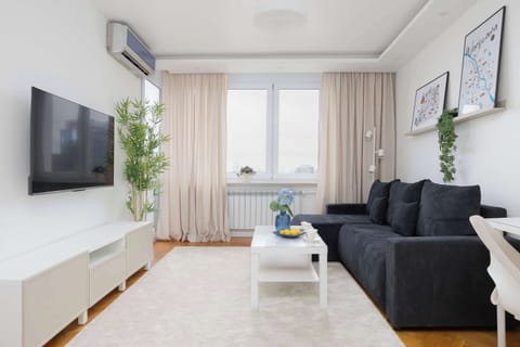 A modern, bright living room features a dark gray sofa, a white coffee table standing on a light-colored rug, and a large television mounted on the wall. A tall window allows plenty of natural light to fill the space.