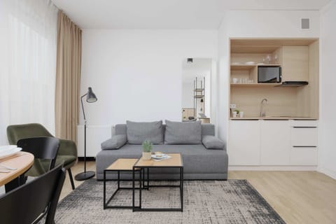 A compact studio apartment with a gray sofa, a small coffee table, and a kitchenette with light wood cabinets along the back wall. Soft neutral tones and minimal decor create a modern atmosphere.