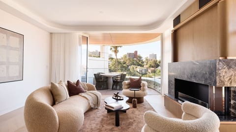 A sophisticated neutral-toned living room with curved boucle furniture, a marble fireplace, and floor-to-ceiling glass doors opening to a sunny balcony.