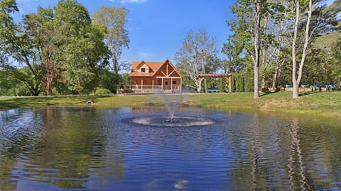 A log cabin with a copper roof overlooks a pond with a central water fountain and surrounding green trees.