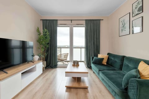 A modern living room features a dark green sofa adorned with yellow and green pillows, set against a light wood floor. A large window allows natural light to fill the space, highlighting the clean and minimalist decor.