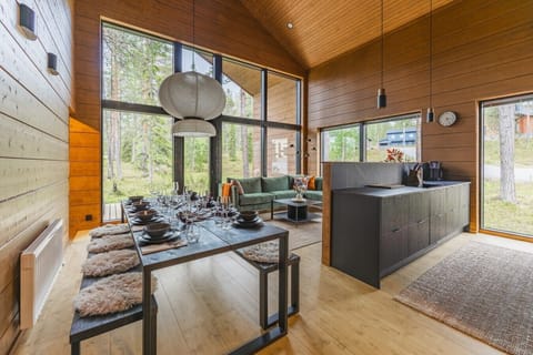 The main floor opens in one bright sweep - dining table, green corner sofa and open kitchen under a vaulted timber ceiling, with a full-height window wall onto the pines.