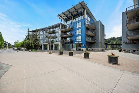 Modern multi-level property featuring contemporary design with striking blue accents and spacious balconies, set in a scenic area surrounded by forested hillsides and open plaza spaces.