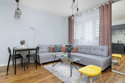 A bright living room with a gray corner sofa, dining table, and large windows. Colorful cushions and modern decor create a cozy atmosphere.