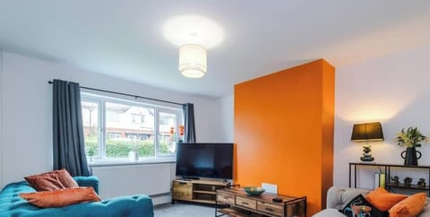 A bright and inviting modern living room designed for comfort and style. This spacious lounge features a bold orange accent wall, a plush teal sofa, and a cozy cream sofa with matching orange cushions that add a warm, vibrant feel.
