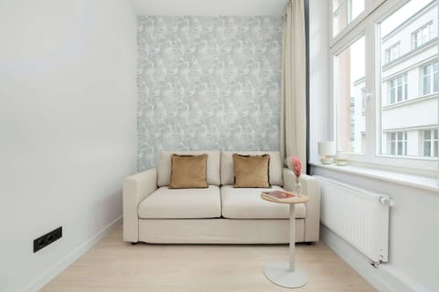 A bright studio apartment featuring a comfortable fold-out sofa, soft neutral tones, and large windows with plenty of natural light.