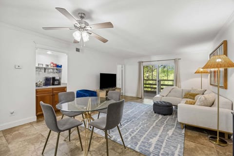 Bright open-concept living space with modern furnishings, a cozy sectional, smart TV, dining for four, and a convenient coffee bar. Sliding glass doors lead to a private balcony with peaceful greenery views.