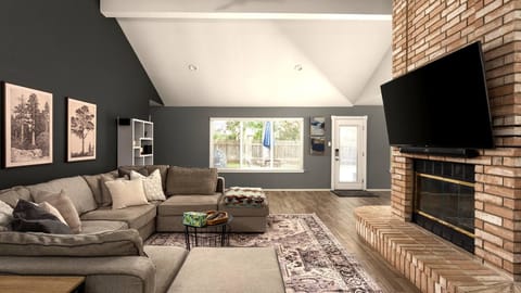 Vaulted-ceiling living room with a large sectional sofa, patterned area rug, wall-mounted TV, and a charming brick fireplace — a spacious and character-filled space that feels warm and welcoming from the moment you walk in.