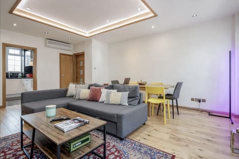 Spacious open-plan living room with air conditioning, L-shaped sofa and dining table seating 6 — coffered ceiling with ambient lighting and direct kitchen access, moments from Oxford Street.