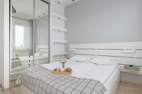 A bright bedroom with a double bed, white shelving units, and large windows covered with sheer curtains. Soft gray tones and modern lighting create a calm and elegant atmosphere.