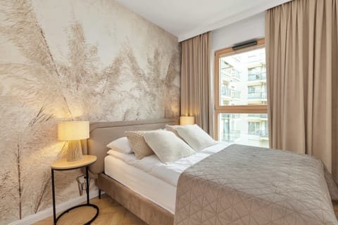 A cozy and bright bedroom featuring a large double bed with plush pillows and a soft blanket. The wall behind the headboard is decorated with a stylish, light-colored botanical wallpaper, and a large window lets in natural light.