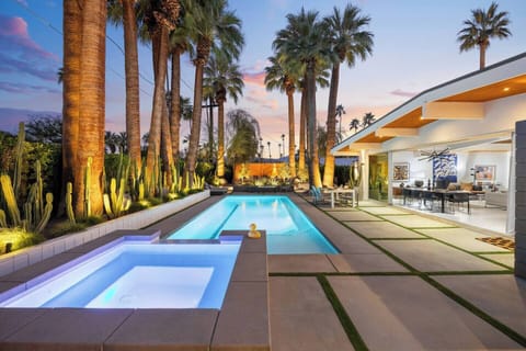 Resort-style backyard at sunset with a heated pool, spa, glowing palms, and warm interior lighting.