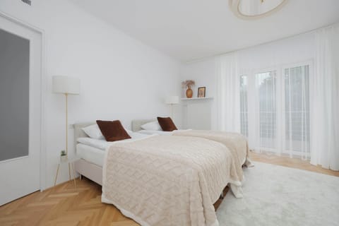 A bright bedroom with a large bed covered in soft beige bedding and decorative pillows. Natural light from the balcony doors creates a calm and airy atmosphere.