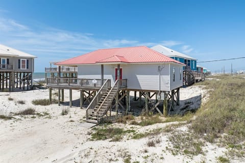 Beachfront cottage on stilts with red metal roof, surrounded by sand dunes and coastal vegetation near the shore.