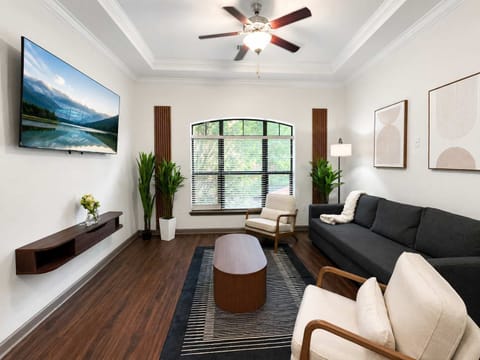Bright, modern living room with high ceilings, wall-mounted TV, large arched window, dark sofa, accent chairs, and warm wood finishes.