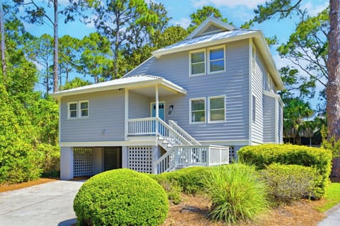 Coastal wood siding and a crisp white porch stand bright against a backdrop of towering pines. This sun-drenched cottage offers a cheerful sky-blue door and lush greenery, creating a peaceful scene for your next getaway.