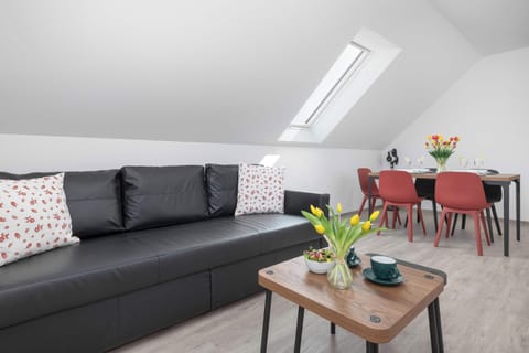 A spacious attic living room is furnished with a sleek black leather sofa and a wooden coffee table displaying a vase of fresh yellow tulips. In the background, a dining table paired with red chairs stands beneath a slanted ceiling with a skylight.