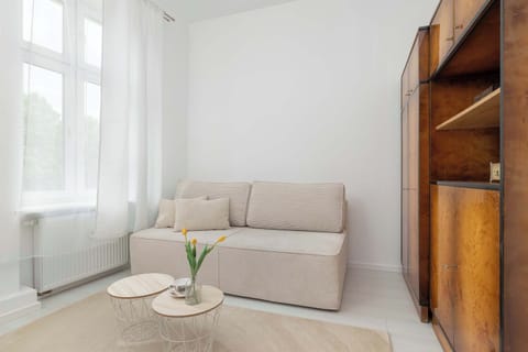 A bright bedroom corner featuring a beige sofa, a small coffee table, and large windows allowing plenty of natural light. Wooden shelving units add warmth and storage to the minimalist interior.