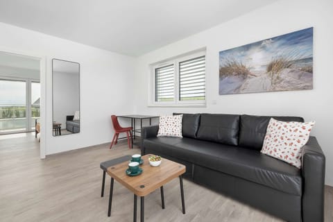 A spacious, sunlit living room features a sleek black leather sofa decorated with patterned cushions, positioned across from a wooden coffee table. A large sea-themed canvas print adorns the main wall, adding a serene element to the space.