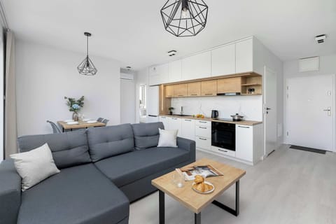 A modern open-plan living room with a gray sectional sofa, wooden coffee table, and compact kitchen in the background. Bright white walls and large windows create a spacious atmosphere.