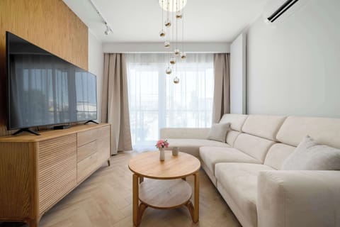 A bright living room with a beige sofa and modern wooden furniture creates a cozy atmosphere. Large windows bring plenty of natural light into the apartment.