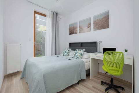 A bright bedroom with a neatly made bed, wall art, and a lime-green office chair beside a compact desk. Large windows allow natural light to fill the minimalist interior.