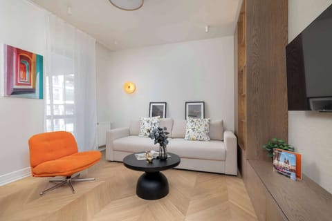 A bright and modern living room offers a comfortable space to relax. The interior features an orange armchair, a sofa, a coffee table, and a wall-mounted TV, creating a cozy atmosphere.
