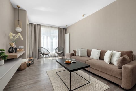 A contemporary living room is decorated in soft beige and brown tones, featuring a large plush sofa with white throw pillows. A black metal coffee table rests on a textured cream rug over wood-look flooring.