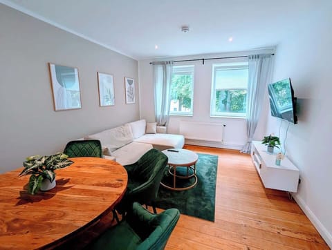 Wohnzimmer mit rundem Esstisch aus Eiche, grünen Samtstühlen, weißem Ecksofa und Blick auf die Bäume vor dem Fenster.