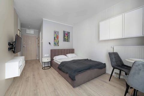 A modern studio apartment featuring a bed, compact kitchenette, and small workspace arranged within one open space. Bright walls and light wood flooring create a clean contemporary atmosphere.