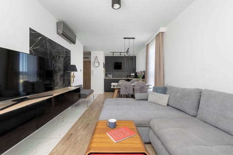A bright living room with a gray sofa and a wall-mounted TV. The open space connects to the dining and kitchen area.
