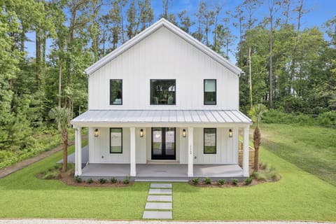 Modern farmhouse rental nestled in a serene wooded setting, featuring clean white exteriors, a welcoming front porch, and lush landscaping for a peaceful getaway.