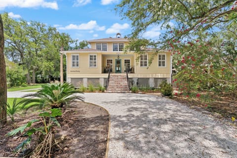 Charming yellow coastal home nestled among mature trees and lush landscaping, featuring inviting front steps and a serene gravel driveway marking the arrival experience.