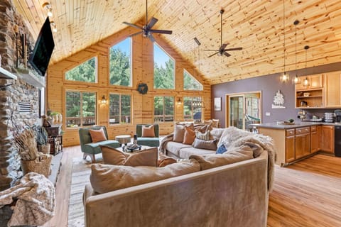 Expansive open-concept living area with soaring wood ceilings, dramatic windows, a stone fireplace, and warm cabin-inspired finishes.