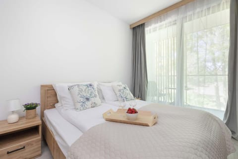 A cozy bedroom with a large double bed, adorned with decorative pillows and rolled-up towels. Light-colored curtains cover the large window, and a wooden nightstand stands next to the bed.