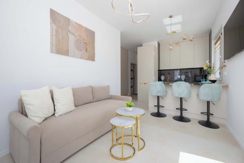A bright living room features a comfortable beige sofa with throw pillows and a modern double coffee table. In the background, an open-plan kitchen with a breakfast bar and blue bar stools is visible.