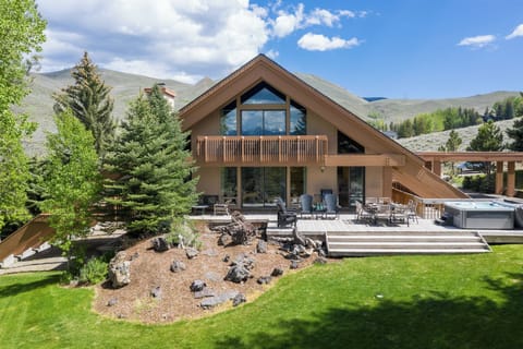 A stunning mountain retreat with expansive patio seating, lush landscaping, and a private hot tub, surrounded by evergreen trees and dramatic alpine peaks.